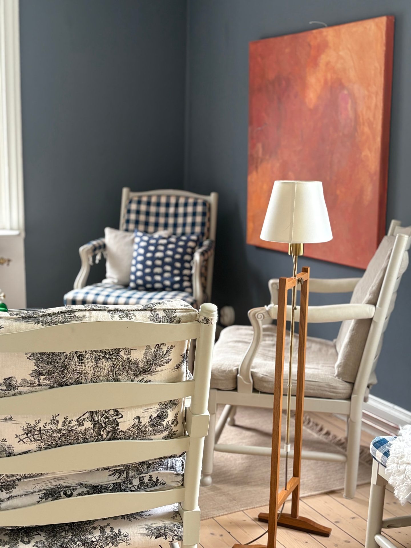 Vardagsrum med väggar målade i kimröksgrå, vita fönsterlister, fåtöljer i toile de Jouy och blåvit rut, golvlampa med cremevit skärm och en tavla i varma toner