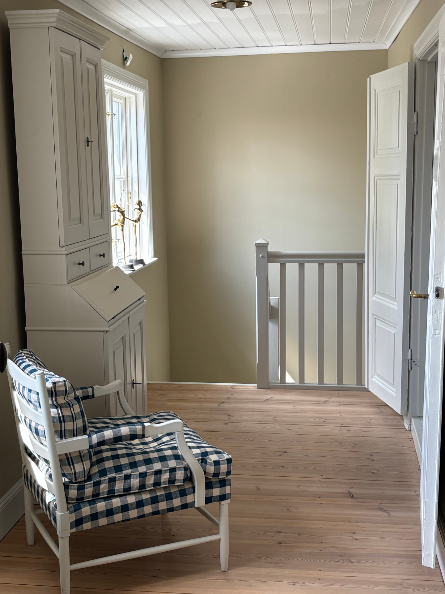 Övre hall med väggar målade i varm beige matt linoljefärg Ax från Ottosson Färgmakeri, vitmålad sekretär, karmstol med rutig dyna, spröjsat fönster med mässingsfigurer på fönsterbrädan, vit spegeldörr och såpat furugolv